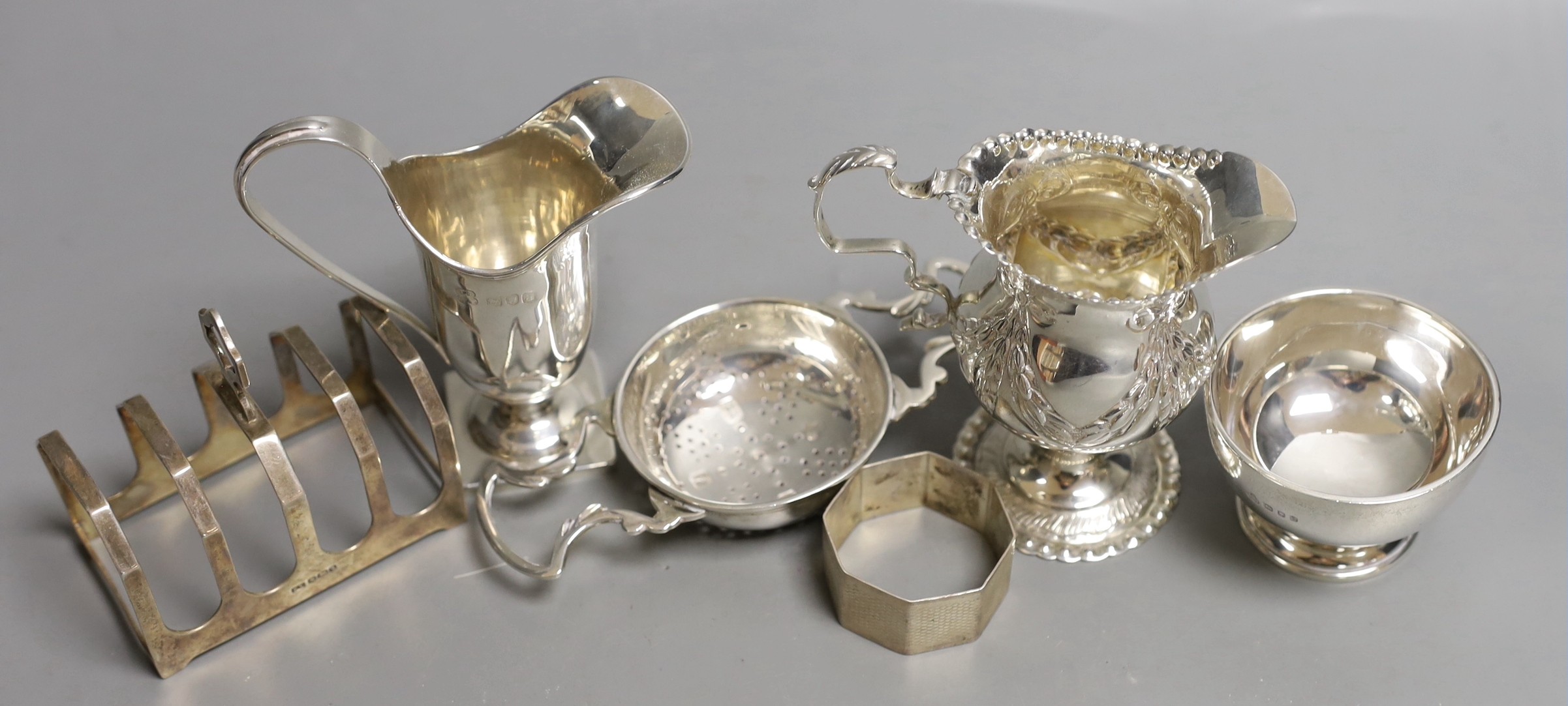 Two late Victorian silver cream jugs, including inverted pear shape, London, 1897, 96mm, a later small silver toastrack, a silver napkin ring and a modern silver tea strainer on stand.